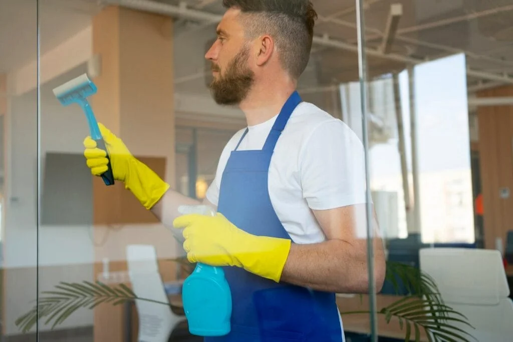 Man in apron and gloves performing window cleaning on a glass window with a squeegee and spray bottle.