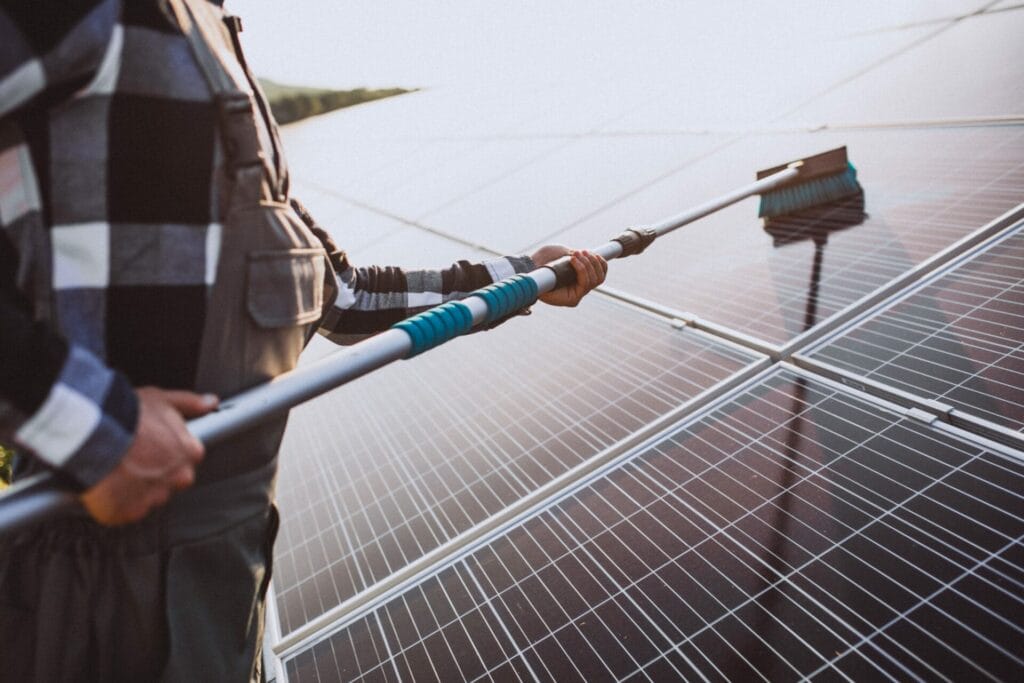 A Person In A Plaid Shirt And Overalls Performs Solar Panel Cleaning Outdoors, Using A Brush On A Pole To Keep The Panels Spotless.