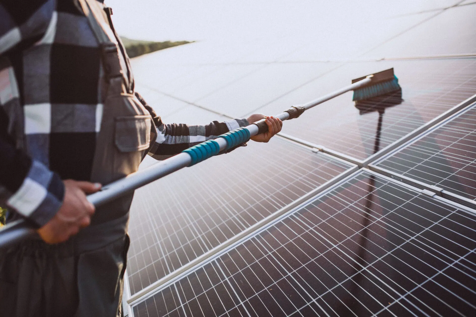 A Person In A Plaid Shirt And Overalls Performs Solar Panel Cleaning Outdoors, Using A Brush On A Pole To Keep The Panels Spotless.